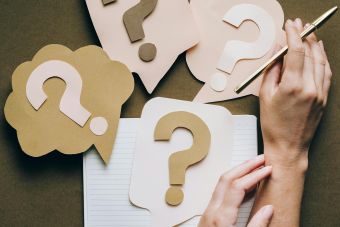Hands holding pen next to paper question marks on notebook, symbolizing inquiry and creativity.